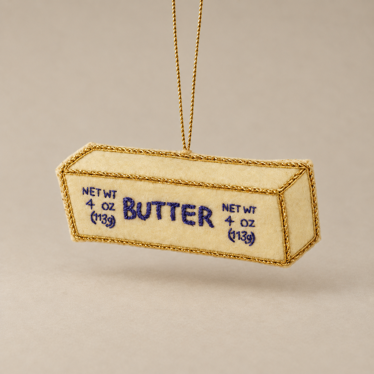 A butter-shaped ornament or decorative piece hanging from a thin gold string, with a cream-colored body, gold braided edges, and blue embroidered text reading 'BUTTER' and 'NET WT 4 oz (13g)'.