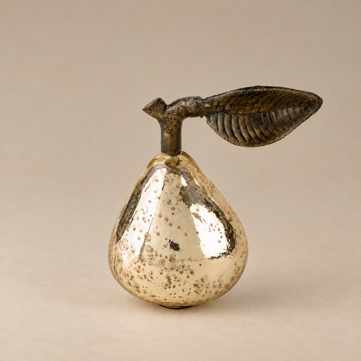 A decorative pear-shaped object with a metallic silver body speckled with gold, topped with a dark brown leaf and stem, set against a plain beige background.