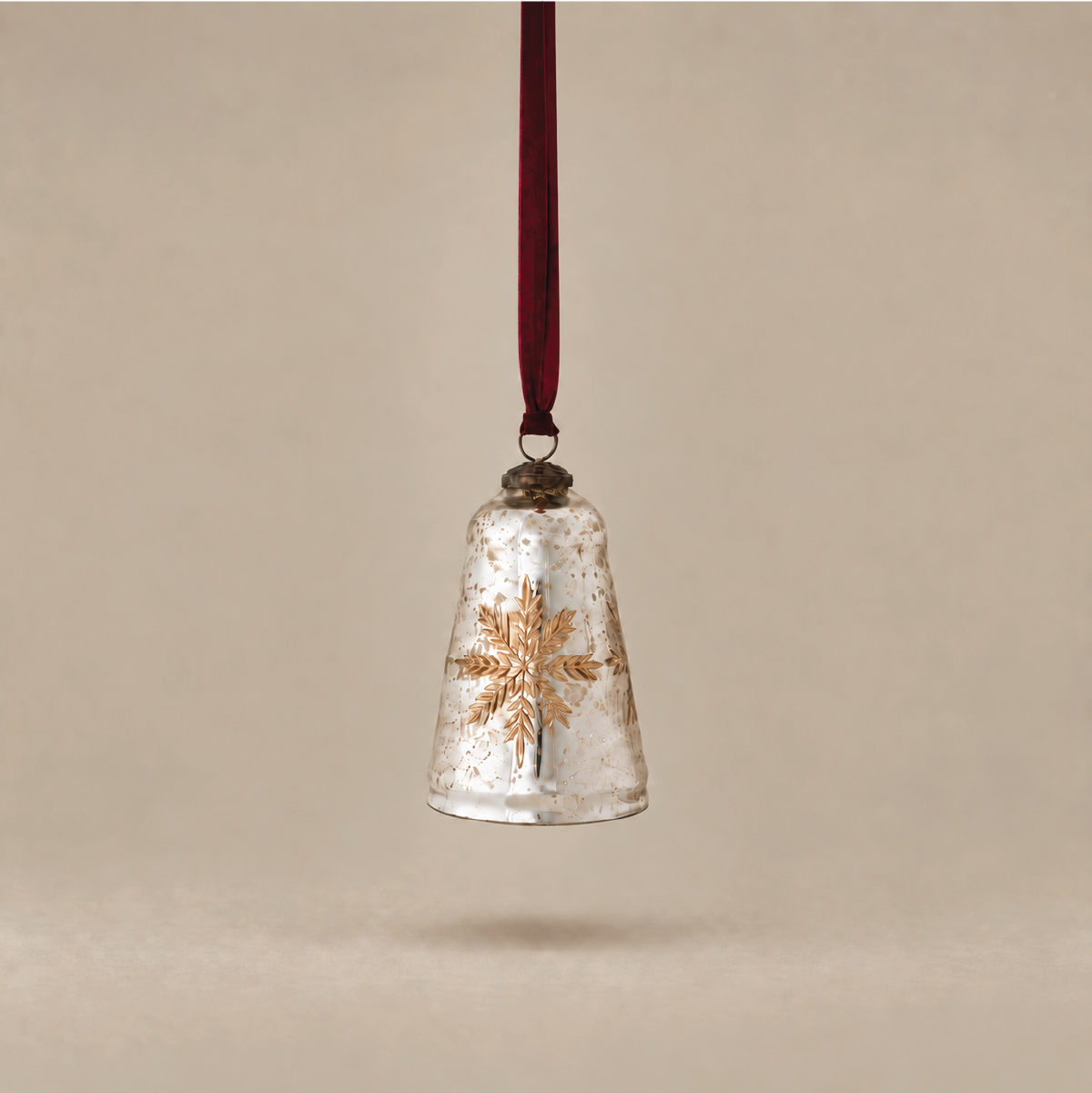 A vintage-style Christmas bell ornament made of clear, textured glass with a metallic gold snowflake design, hanging from a dark red ribbon against a warm beige background.