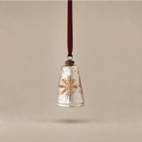 A vintage-style Christmas bell ornament made of clear, textured glass with a metallic gold snowflake design, hanging from a dark red ribbon against a warm beige background.