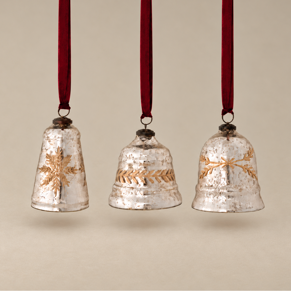 Three vintage-style hanging Christmas bells with a textured silver finish and gold decorative motifs, suspended by dark red ribbons against a warm beige background.