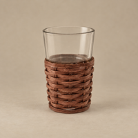 A clear glass tumbler with a brown woven rattan or wicker wrap around the lower half, shown on a plain beige studio background.