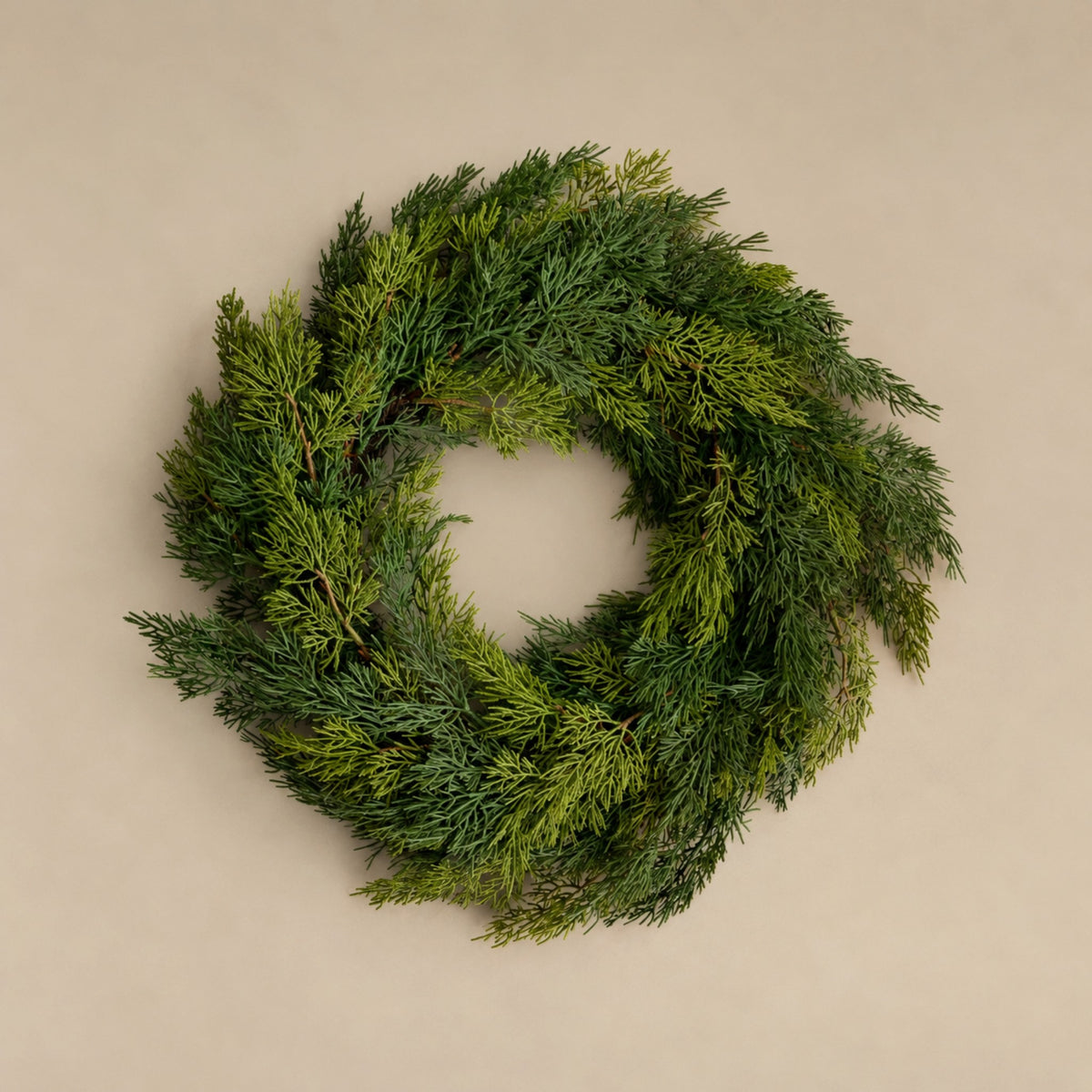 A lush green evergreen wreath made of dense pine or cedar branches, photographed from directly above on a plain beige background.