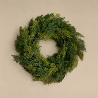 A lush green evergreen wreath made of dense pine or cedar branches, photographed from directly above on a plain beige background.