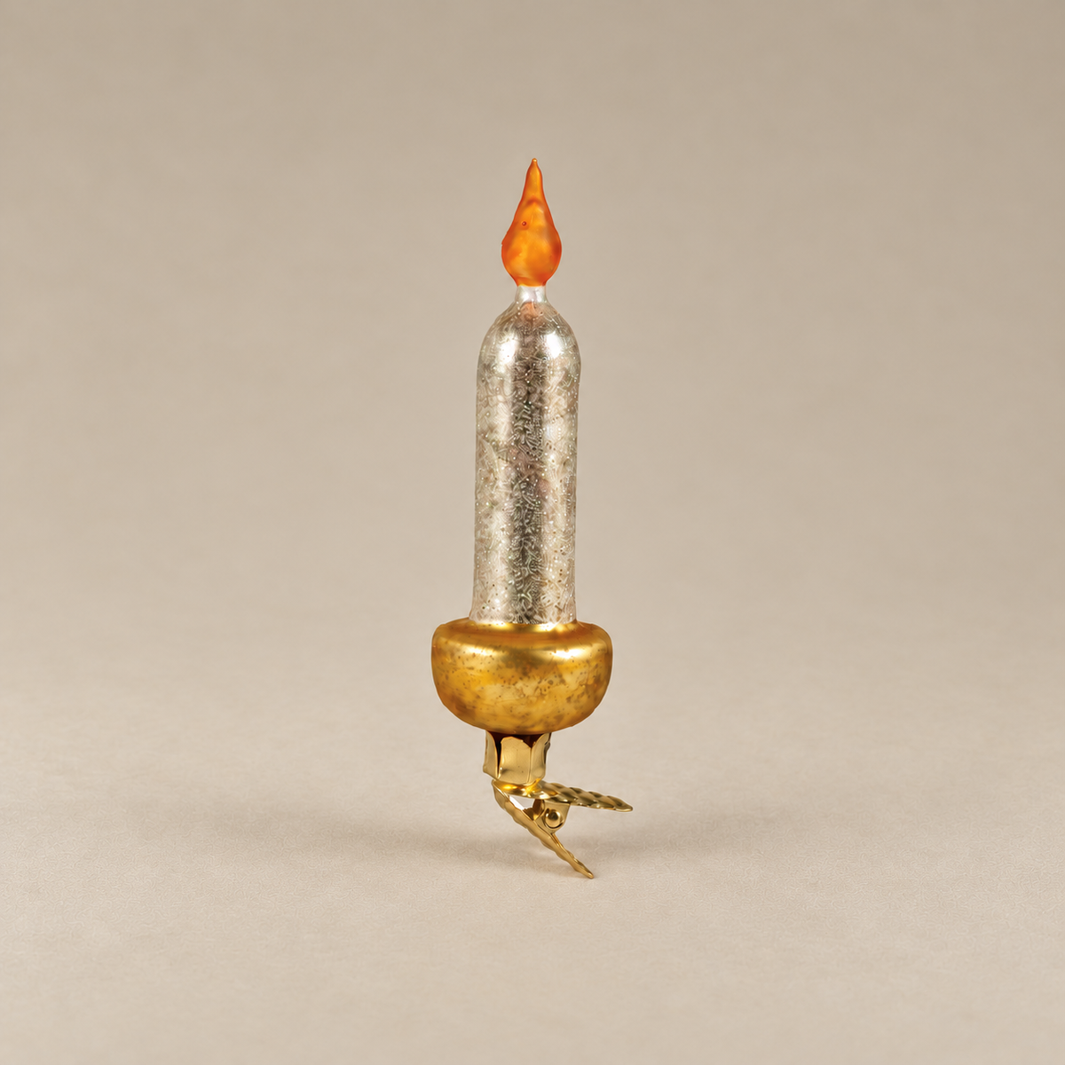 A miniature decorative candle-shaped ornament with a textured silver body, a gold-colored base, and an orange flame tip, set against a solid neutral beige background.