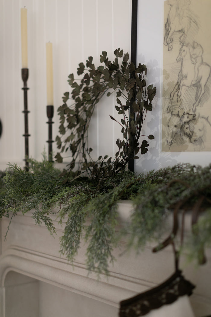Decorative wreath and greenery on a mantelpiece with candles and artwork in the background.